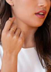 A woman with long brown hair, in a white ribbed top and natural makeup, softly touches her face while showcasing the panacheaccessories Luxe Gold Starburst Stamped Ring on her finger.