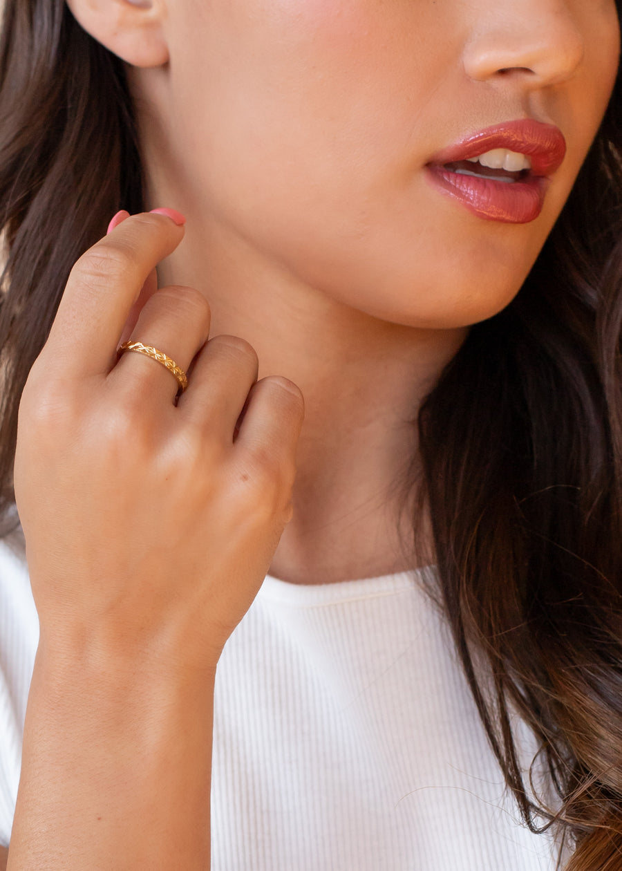 A woman with long brown hair, in a white ribbed top and natural makeup, softly touches her face while showcasing the panacheaccessories Luxe Gold Starburst Stamped Ring on her finger.