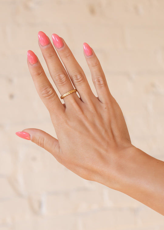 A hand with long pink manicured nails wears the Luxe Gold Stamped Ring by panacheaccessories on the middle finger, set against a light beige background.