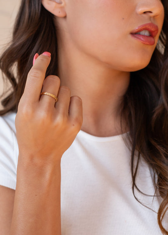 A woman with long brown hair, wearing a white ribbed top, poses with her hand by her chin to showcase the Luxe Gold Stamped Ring by panacheaccessories. Her nails are painted pink and her lips are slightly parted.