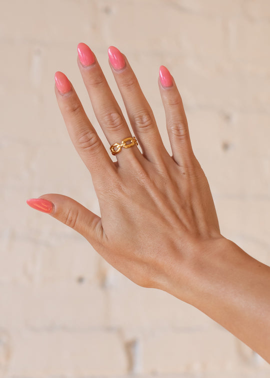 A hand with long, glossy pink nails wears the Luxe Gold Wide Paper Clip Link Ring by panacheaccessories on the middle finger, held up against a blurred light-colored brick wall background.