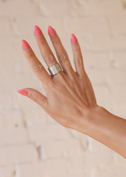 A hand with pink nails is held up against a light wall, showing off the panacheaccessories Luxe Silver Textured Ring on the middle finger. This adjustable statement piece ensures a perfect fit.