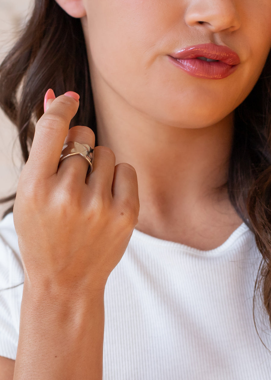 A woman with wavy brown hair and a white top holds her hand near her face, showcasing the Luxe Silver Wide Hammered Ring by panacheaccessories—an eye-catching chunky adjustable stainless steel piece.