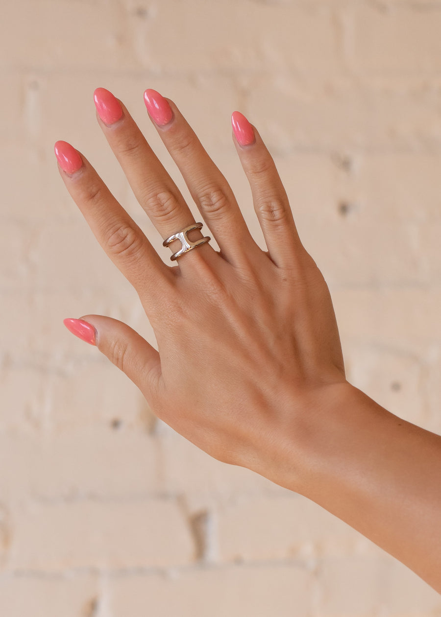 A hand with long, glossy pink nails showcases the panacheaccessories Luxe Silver Double Layer Ring on the middle finger, creating a bold accent against a light brick wall.