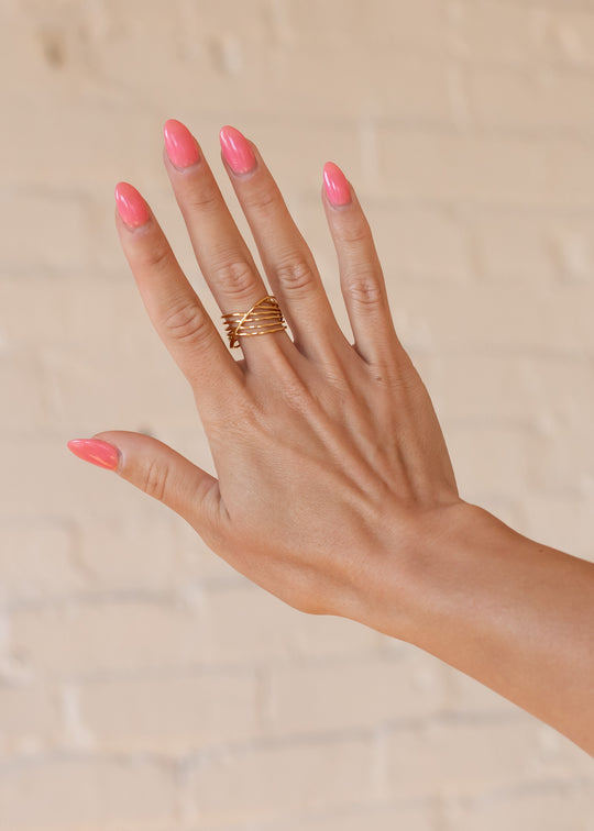 A hand with long, glossy pink nails is raised against a light beige brick wall, showcasing panacheaccessories Luxe Gold Multi-Strand Ring—an elegant, adjustable 18K gold band with intertwined strands for effortless chic.