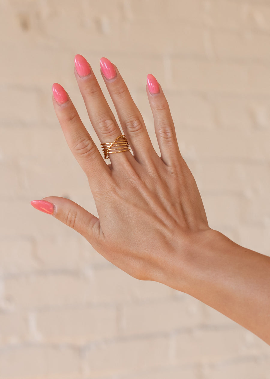 A hand with long, glossy pink nails is raised against a light beige brick wall, showcasing panacheaccessories Luxe Gold Multi-Strand Ring—an elegant, adjustable 18K gold band with intertwined strands for effortless chic.