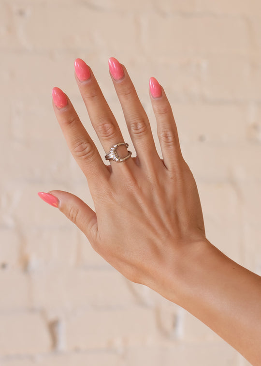 A hand with pink, glossy manicured nails is shown against a light brick wall, wearing the panacheaccessories Luxe Silver Decorative Ring featuring a clear stone on the ring finger.
