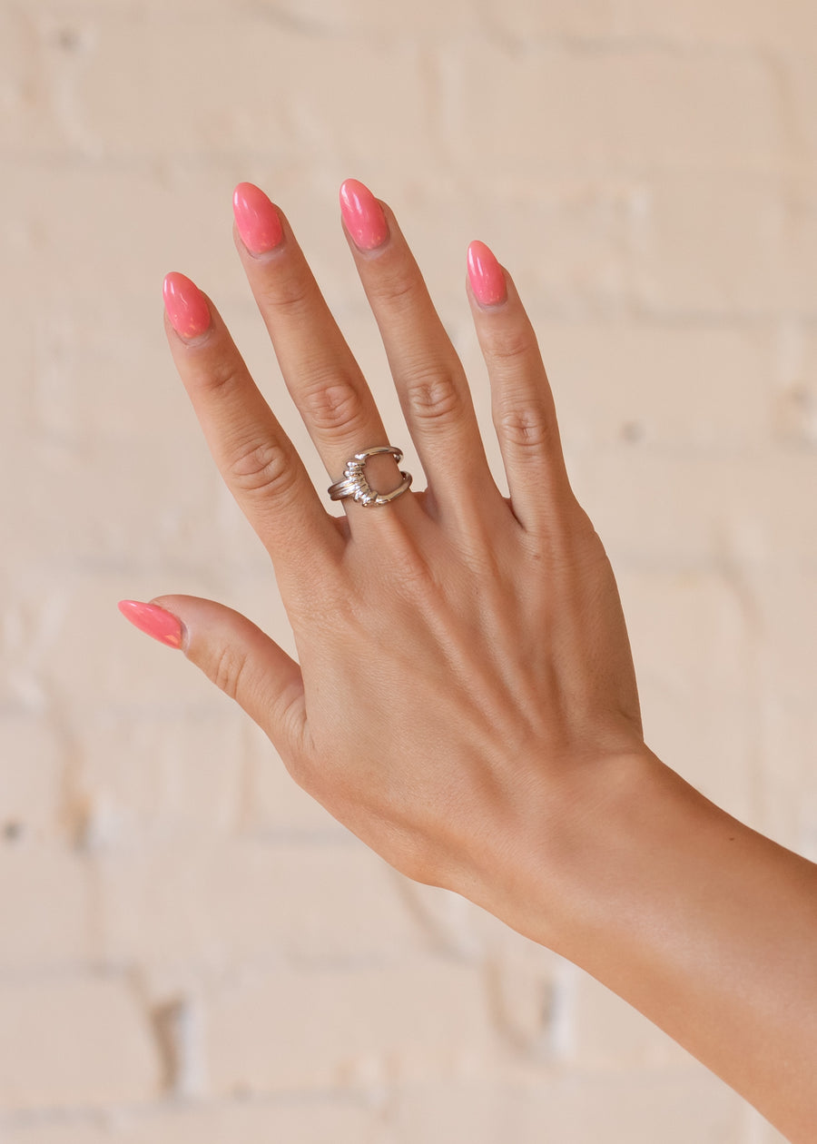 A hand with pink, glossy manicured nails is shown against a light brick wall, wearing the panacheaccessories Luxe Silver Decorative Ring featuring a clear stone on the ring finger.
