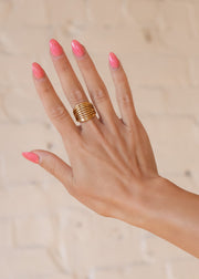 A hand with pink-painted nails showcases panacheaccessories Luxe Gold Wide Multi-Strand Ring, an adjustable 18K gold statement piece, worn on the middle finger against a softly blurred background.