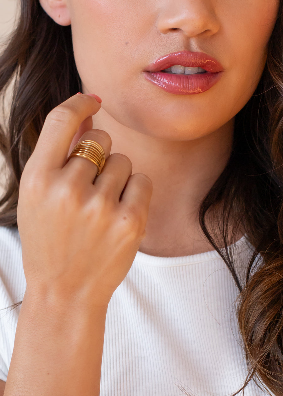 A woman with wavy brown hair and glossy lips touches her face, wearing a white ribbed top and the Luxe Gold Wide Multi-Strand Ring by panacheaccessories stacked on her fingers.