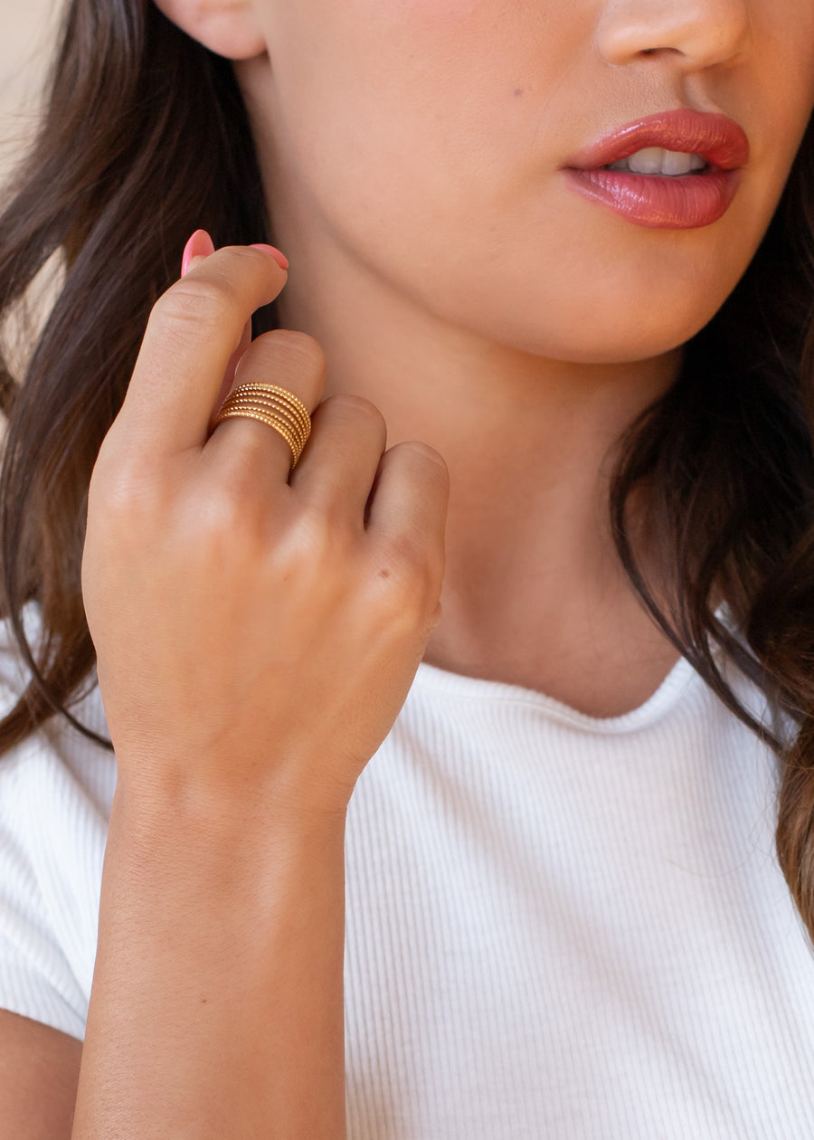 A woman in a white ribbed shirt touches her lips with her left hand, wearing panacheaccessories’ Luxe Gold Dainty Multi-Strand Ring. Her wavy brown hair frames her face, and she sports natural makeup with pink lipstick.