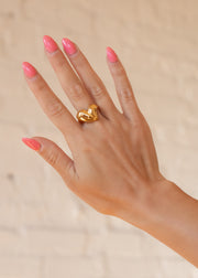 A hand with pink, almond-shaped nails showcases the Luxe Gold Heart Shaped Ring by panacheaccessories against a light beige brick wall background.