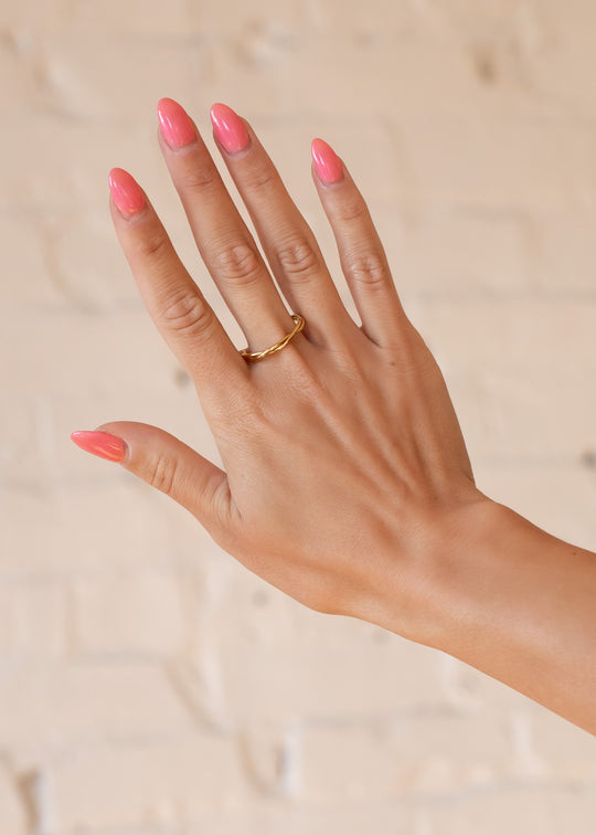 A hand with a glossy pink manicure showcases panacheaccessories Luxe Gold Thin Twisted Ring in 18K gold on the middle finger, set against a light beige brick wall.