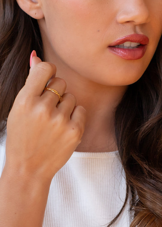 A woman with long brown hair wears a white top and the Luxe Gold Thin Twisted Ring by panacheaccessories, holding her hand near her face. The photo focuses on her natural makeup and glossy pink lips, showing only the lower half of her face.