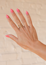 A hand with long, glossy pink nails wears the panacheaccessories Luxe Gold LOVE Ring, adjustable and crafted in 18K gold, across two fingers against a softly blurred beige brick background.
