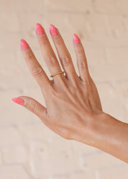 A hand with pink, almond-shaped nails wears the panacheaccessories Luxe Gold Thin Textured Ring on the middle finger, posed against a light beige background.