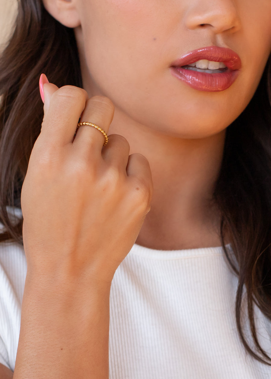 A woman with long brown hair and glossy pink lips wears a white ribbed top, highlighting the Luxe Gold Thin Textured Ring by panacheaccessories on her finger.
