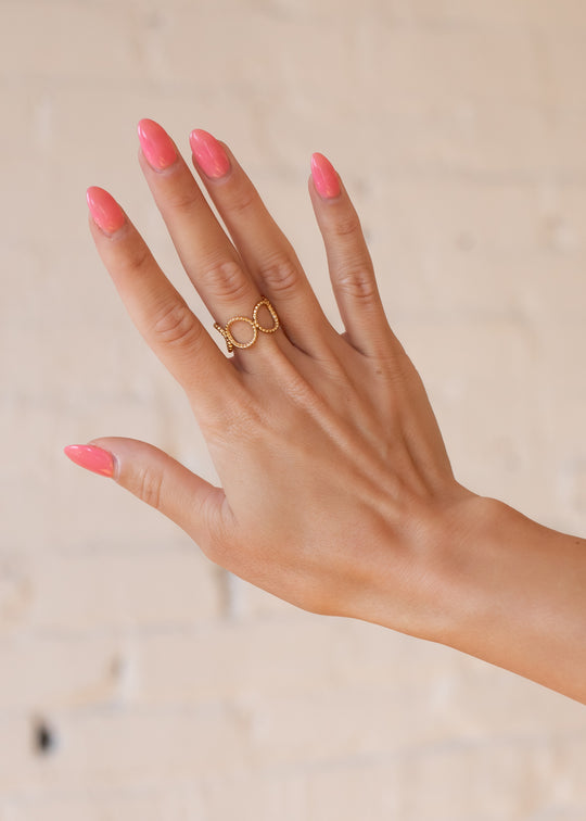 A hand with long, glossy pink nails is shown against a soft background. The middle finger wears panacheaccessories Luxe Gold Open Circles Ring, an adjustable 18K gold piece featuring an elegant open circle design.