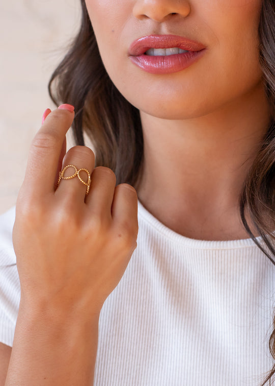 A woman with wavy brown hair wears a white top and the Luxe Gold Open Circles Ring by panacheaccessories, gently touching her face. Her light pink nails, hand, and the lower half of her face are visible.