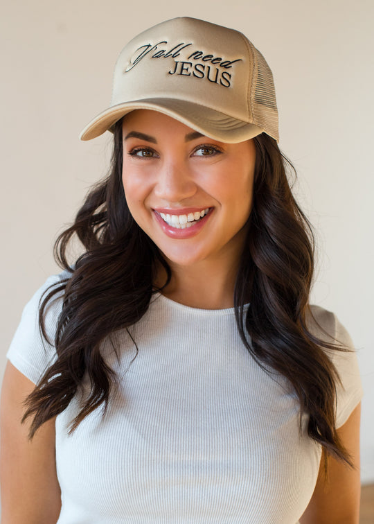 A smiling woman with long dark hair wears a white ribbed short-sleeve shirt and the panacheaccessories Y'all Need Jesus Trucker Hat, which features an adjustable snapback, standing against a plain light background.