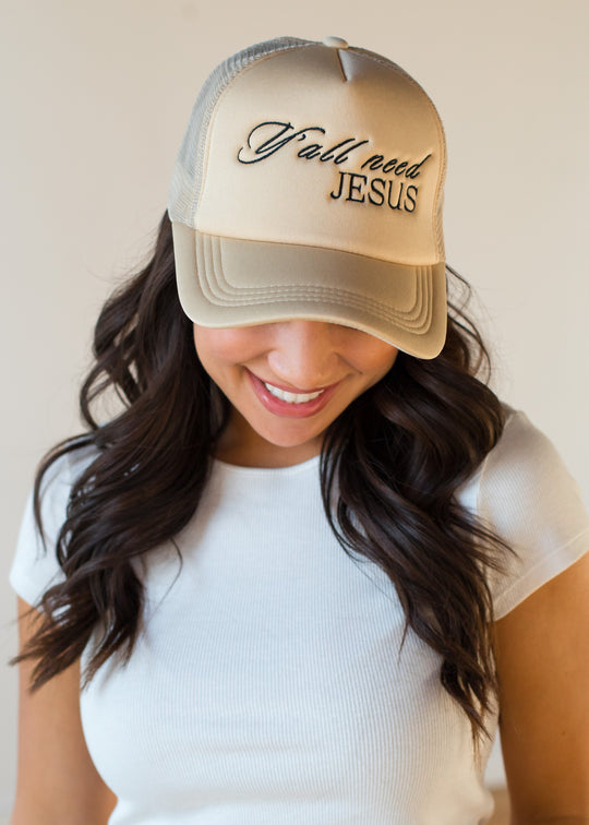 A woman with long dark hair smiles while looking down, wearing the panacheaccessories Y'all Need Jesus Trucker Hat in beige with an adjustable snapback, paired with a white ribbed short-sleeve top against a plain light background.