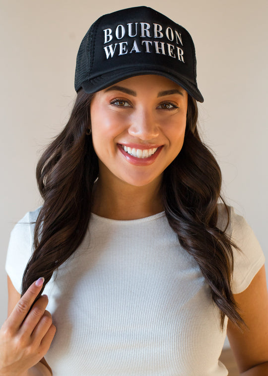 A smiling woman with long dark hair models the panacheaccessories Bourbon Weather Trucker Hat - Black and a short-sleeved white shirt, posing against a plain background.