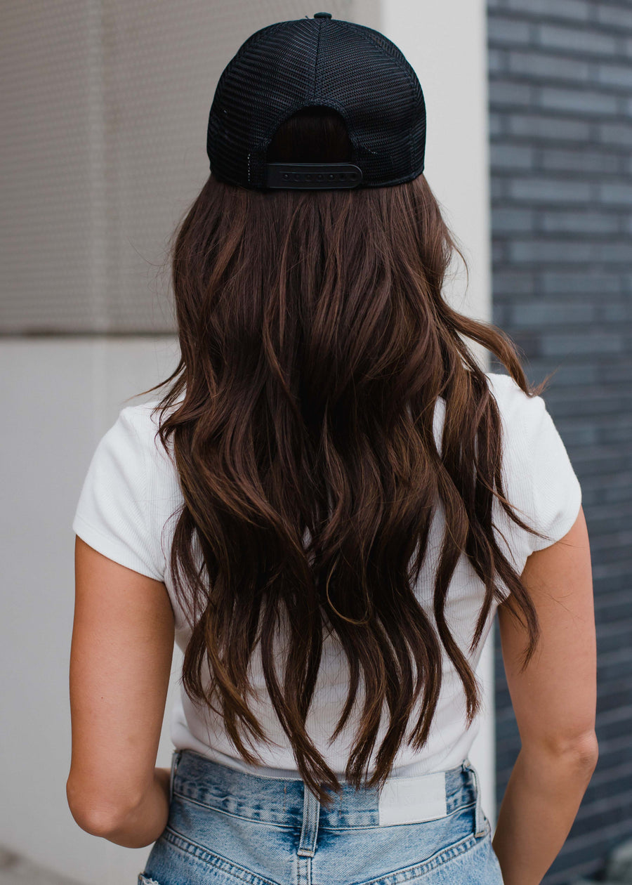 Wearing a panacheaccessories Pink Floyd Trucker Hat backwards, a person with long wavy brown hair, a white short-sleeve shirt, and light blue jeans stands with their back to the camera by a modern building.