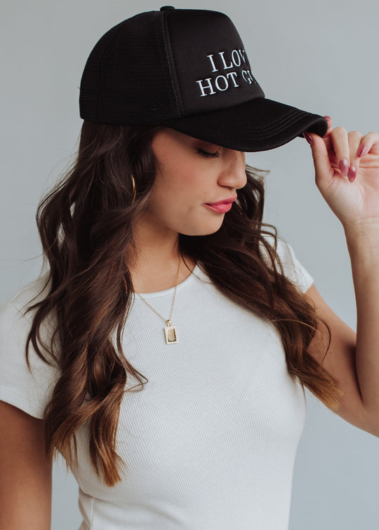 A woman with long brown hair wears a white shirt and a panacheaccessories I Love Hot Guys Trucker Hat, tilting her head down as she holds the brim. She accessorizes with a gold necklace and hoop earrings.