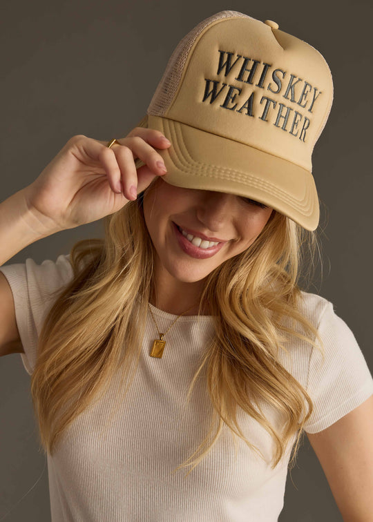 A smiling woman with long blonde hair wears the panacheaccessories Whiskey Weather Trucker Hat in Tan & Gray, paired with a ribbed beige short-sleeve top and gold necklace. She touches the hat’s brim and looks downward.