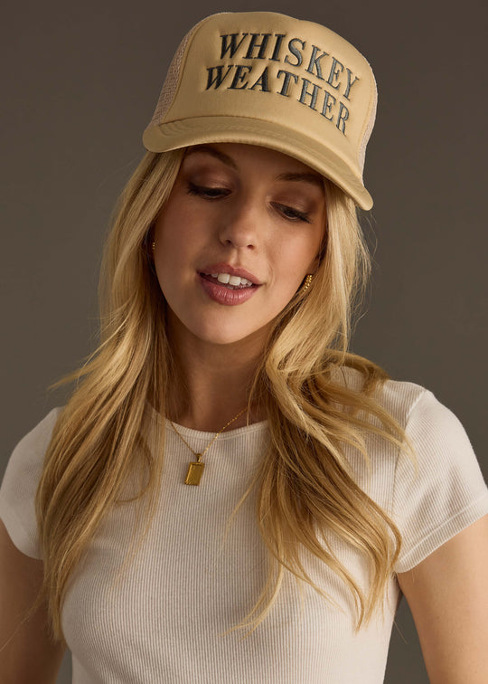 A woman with long blonde hair wears the panacheaccessories Whiskey Weather Trucker Hat in tan & gray, featuring a snapback closure, and a white ribbed short-sleeve top. She looks down with a slight smile against a neutral background.