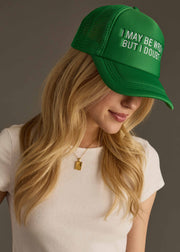 A woman with long blonde hair wears the panacheaccessories I May Be Wrong Trucker Hat, an adjustable green cap, and a white short-sleeve top. She looks down with a slight smile.