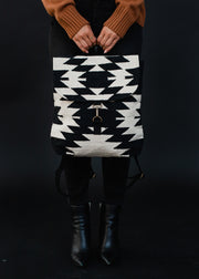 A person in a brown sweater, black pants, and black boots holds the Camilla Backpack by Panache Apparel Collection, featuring adjustable straps, in front of them against a dark background.