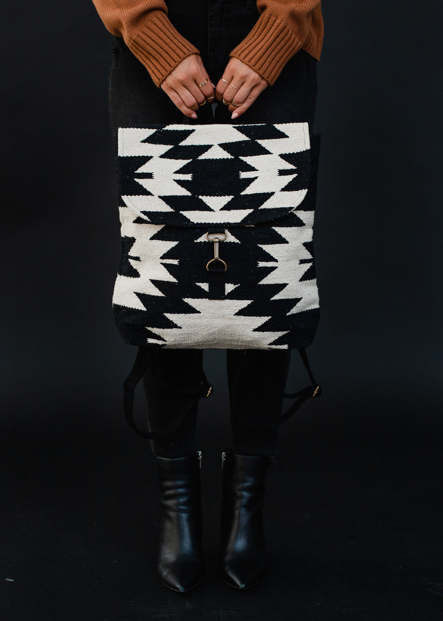 A person in a brown sweater, black pants, and black boots holds the Camilla Backpack by Panache Apparel Collection, featuring adjustable straps, in front of them against a dark background.