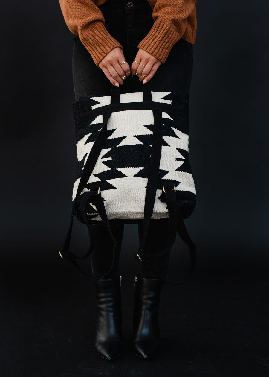 A person dressed in black jeans, black boots, and a brown sweater holds the Camilla Backpack from Panache Apparel Collection, featuring adjustable straps, against a black background.