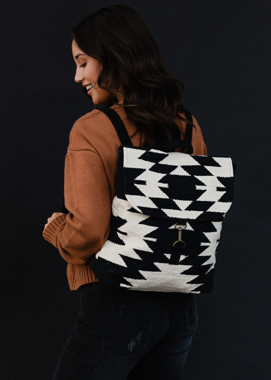 A woman in a brown sweater and black jeans smiles while posing with the Camilla Backpack by Panache Apparel Collection, which features adjustable straps, against a dark background.