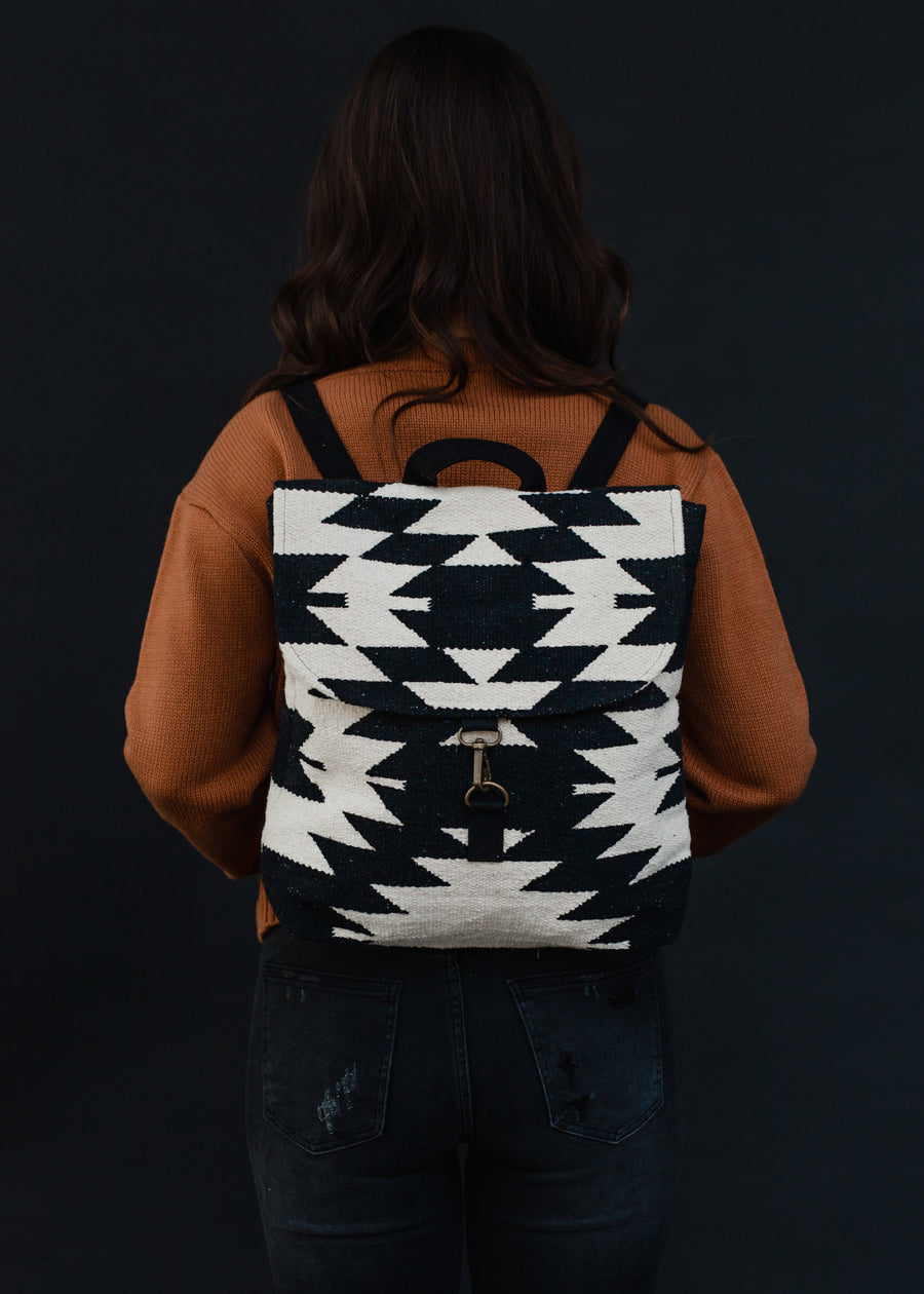 A person with long brown hair, dressed in a brown sweater and dark jeans, stands against a dark background showcasing the Camilla Backpack by Panache Apparel Collection, featuring adjustable straps on their back.