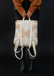 A person in a brown sweater, black jeans, and black boots holds the Celeste Backpack by panacheaccessories, featuring adjustable straps and a beige and white pattern, standing against a black background.