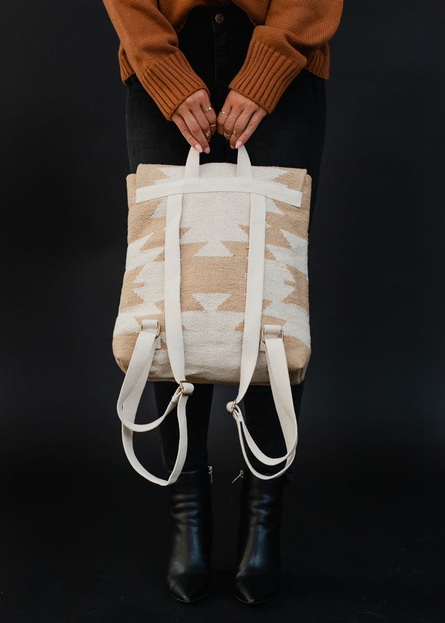 A person in a brown sweater, black jeans, and black boots holds the Celeste Backpack by panacheaccessories, featuring adjustable straps and a beige and white pattern, standing against a black background.