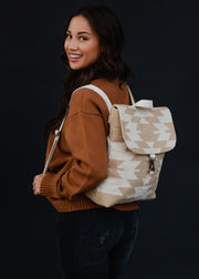 A woman with long dark hair in a brown sweater and black jeans smiles over her shoulder, carrying the Celeste Backpack by panacheaccessories, featuring adjustable straps and a clasp closure against a dark background.