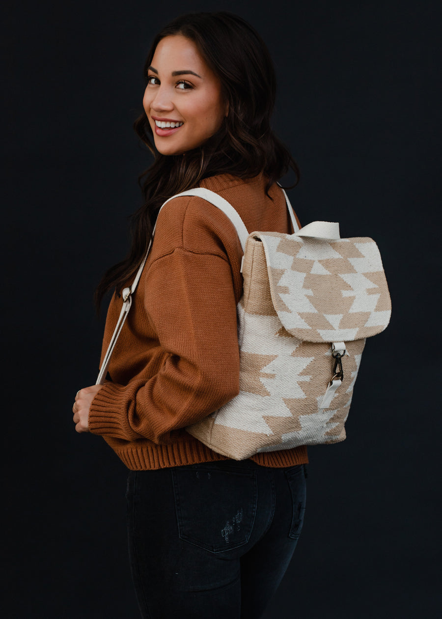 A woman with long dark hair in a brown sweater and black jeans smiles over her shoulder, carrying the Celeste Backpack by panacheaccessories, featuring adjustable straps and a clasp closure against a dark background.