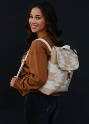 A smiling woman with long brown hair wears a brown sweater, black jeans, and carries the panacheaccessories Celeste Backpack with adjustable straps, posing against a plain dark background.