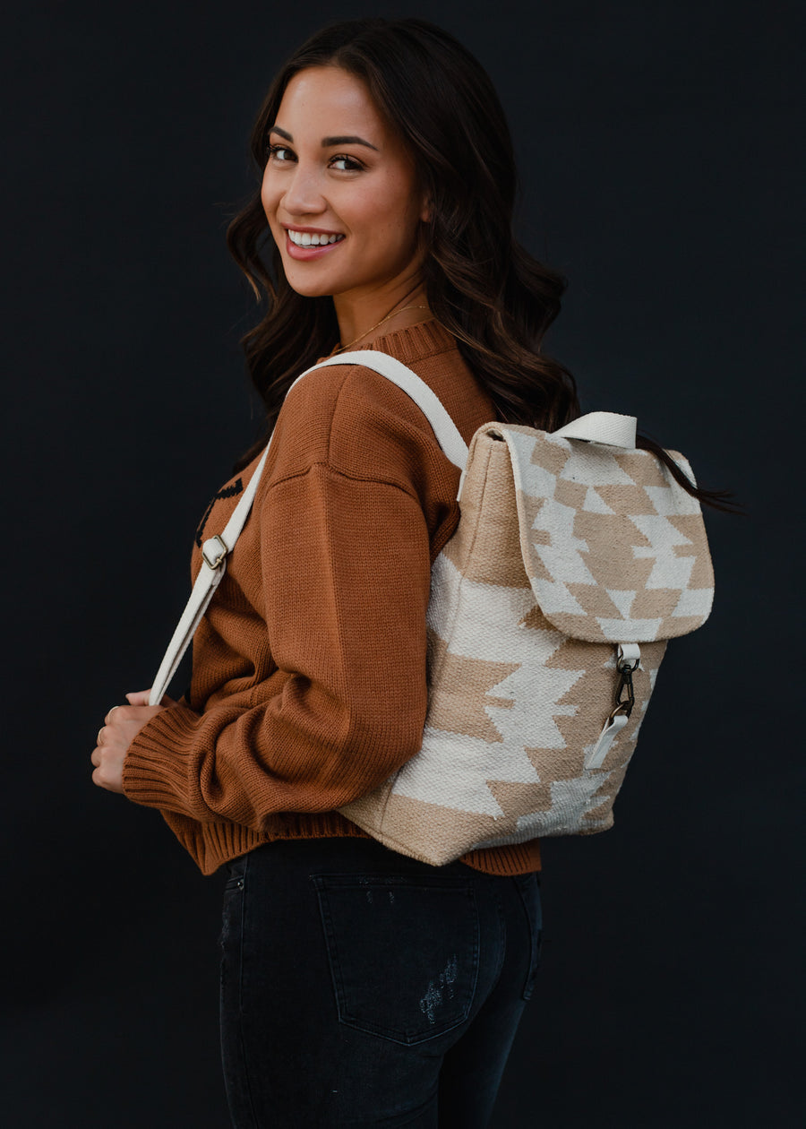 A smiling woman with long brown hair wears a brown sweater, black jeans, and carries the panacheaccessories Celeste Backpack with adjustable straps, posing against a plain dark background.