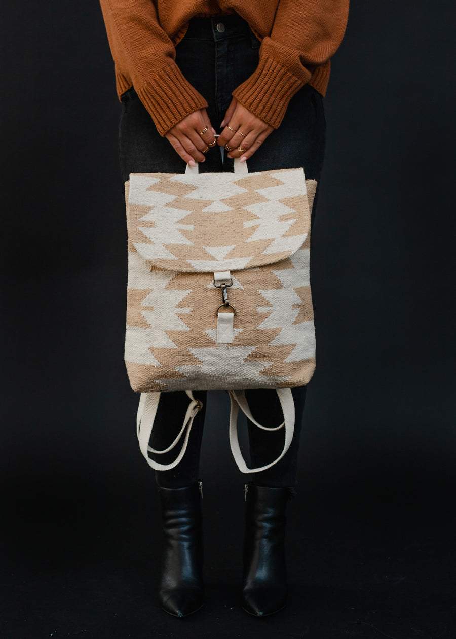 A person in a brown sweater and black pants holds the Celeste Backpack by panacheaccessories—beige and white with geometric designs, clasp closure, and adjustable straps—standing against a black background.