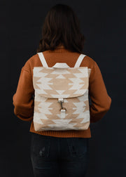 A person with wavy brown hair in a rust sweater and dark jeans faces away, carrying the panacheaccessories Celeste Backpack, which features adjustable straps and a beige and white pattern on a black background.