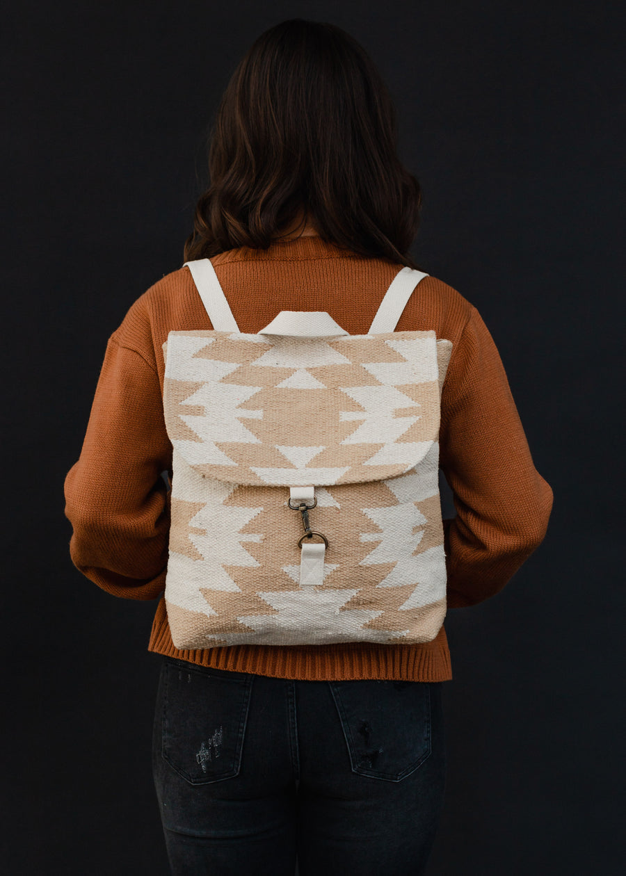 A person with wavy brown hair in a rust sweater and dark jeans faces away, carrying the panacheaccessories Celeste Backpack, which features adjustable straps and a beige and white pattern on a black background.