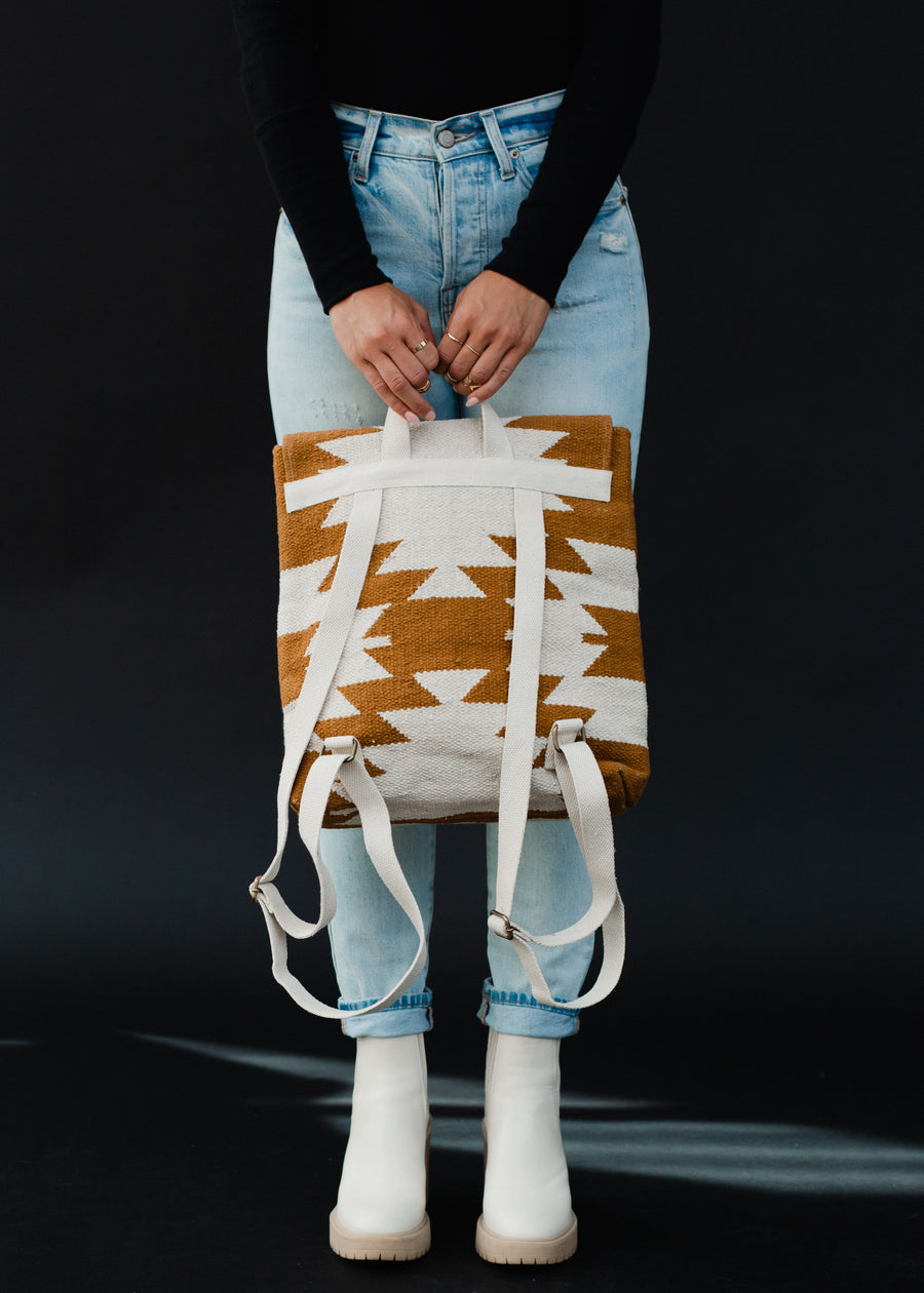 A person in a black top, light blue ripped jeans, and white boots holds the panacheaccessories Inez Backpack—a western-inspired bag with adjustable straps—in front of them against a black background.