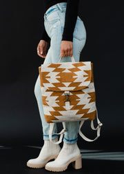 A person in light blue jeans, a black long-sleeve top, and cream heeled boots holds the panacheaccessories Inez Backpack, featuring adjustable straps and a tan and white geometric pattern on a black background.