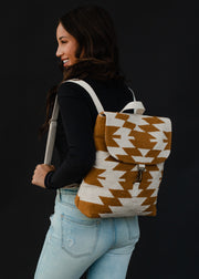 A woman in a black long-sleeve shirt and light blue jeans smiles while carrying the Inez Backpack by panacheaccessories, featuring adjustable straps and a tan and white geometric pattern on a black background.