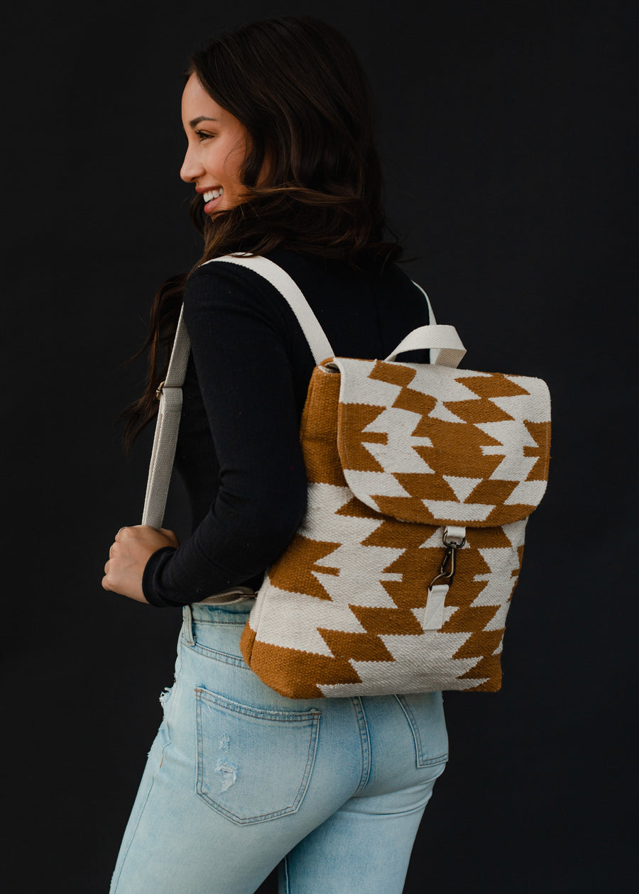 A woman in a black long-sleeve shirt and light blue jeans smiles while carrying the Inez Backpack by panacheaccessories, featuring adjustable straps and a tan and white geometric pattern on a black background.