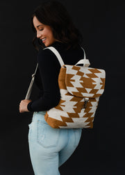 A woman with wavy dark hair smiles as she looks down, wearing a black long-sleeve top, light blue jeans, and the Inez Backpack by panacheaccessories with adjustable straps, set against a black background.